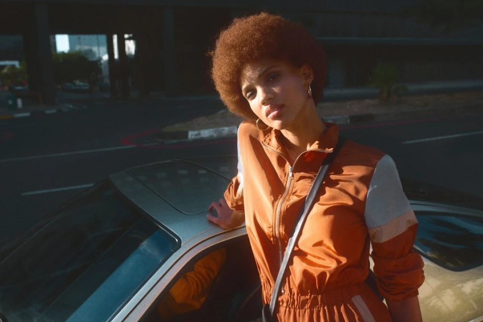 A woman with an afro in an orange tracksuit leans against a silver car on a city street with an overpass in the background.