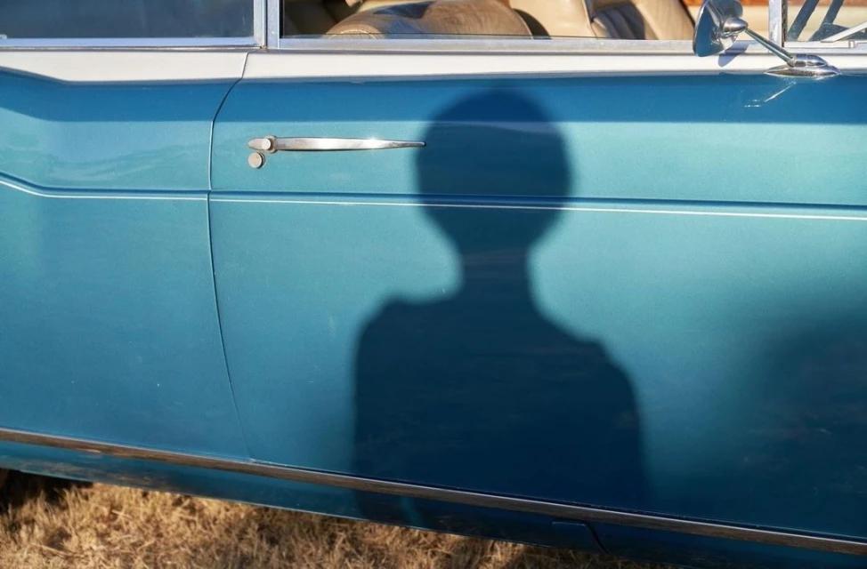 Close-up of a blue car door with a chrome handle and trim, featuring a dark silhouette of a person cast across the surface.
