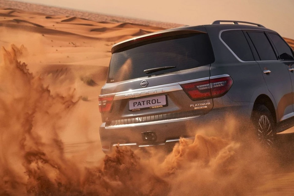 Nissan Patrol SUV driving through a desert landscape with golden sand dunes. The vehicle kicks up sand as it moves, showcasing its rugged design.