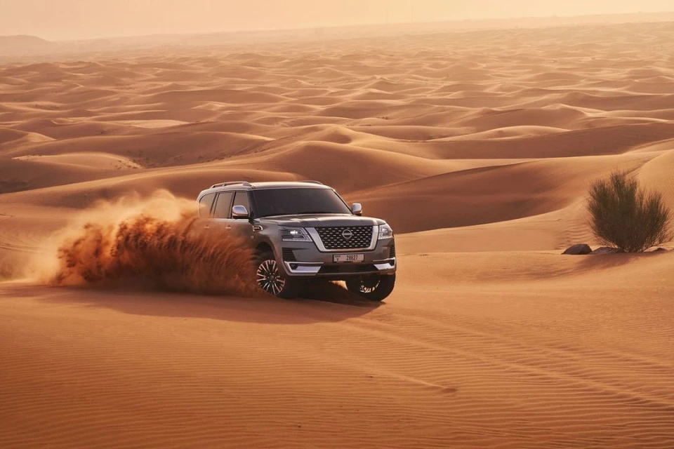 Gray SUV driving across a vast desert landscape with rolling sand dunes under a clear sky, kicking up sand and dust behind the vehicle.