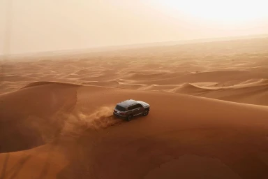 An SUV driving across vast sand dunes kicking up dust, captured from an elevated aerial perspective in golden desert light.