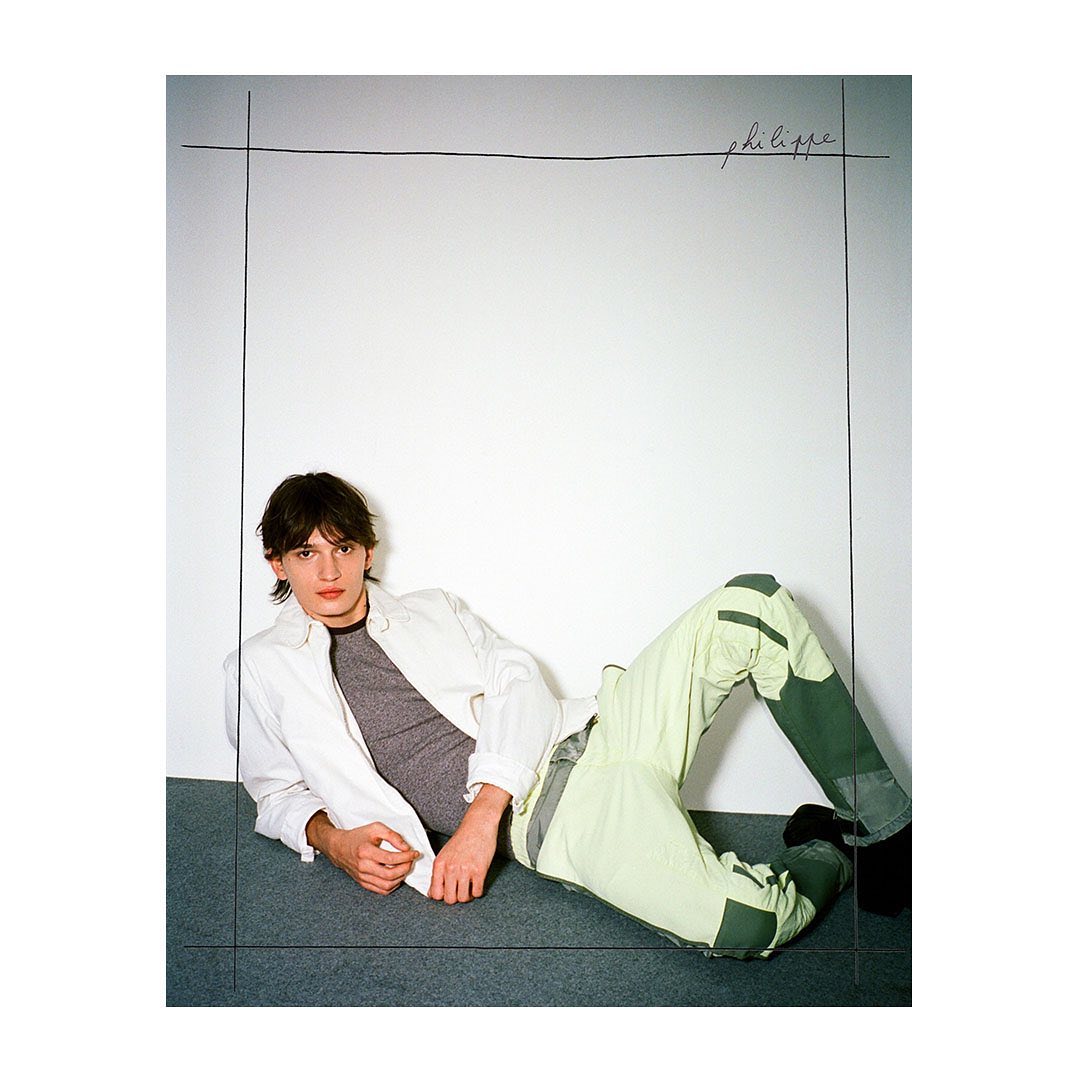 A young adult man is lying on a gray floor in front of a white wall with black framing lines, wearing a gray shirt, light jacket, and pants.