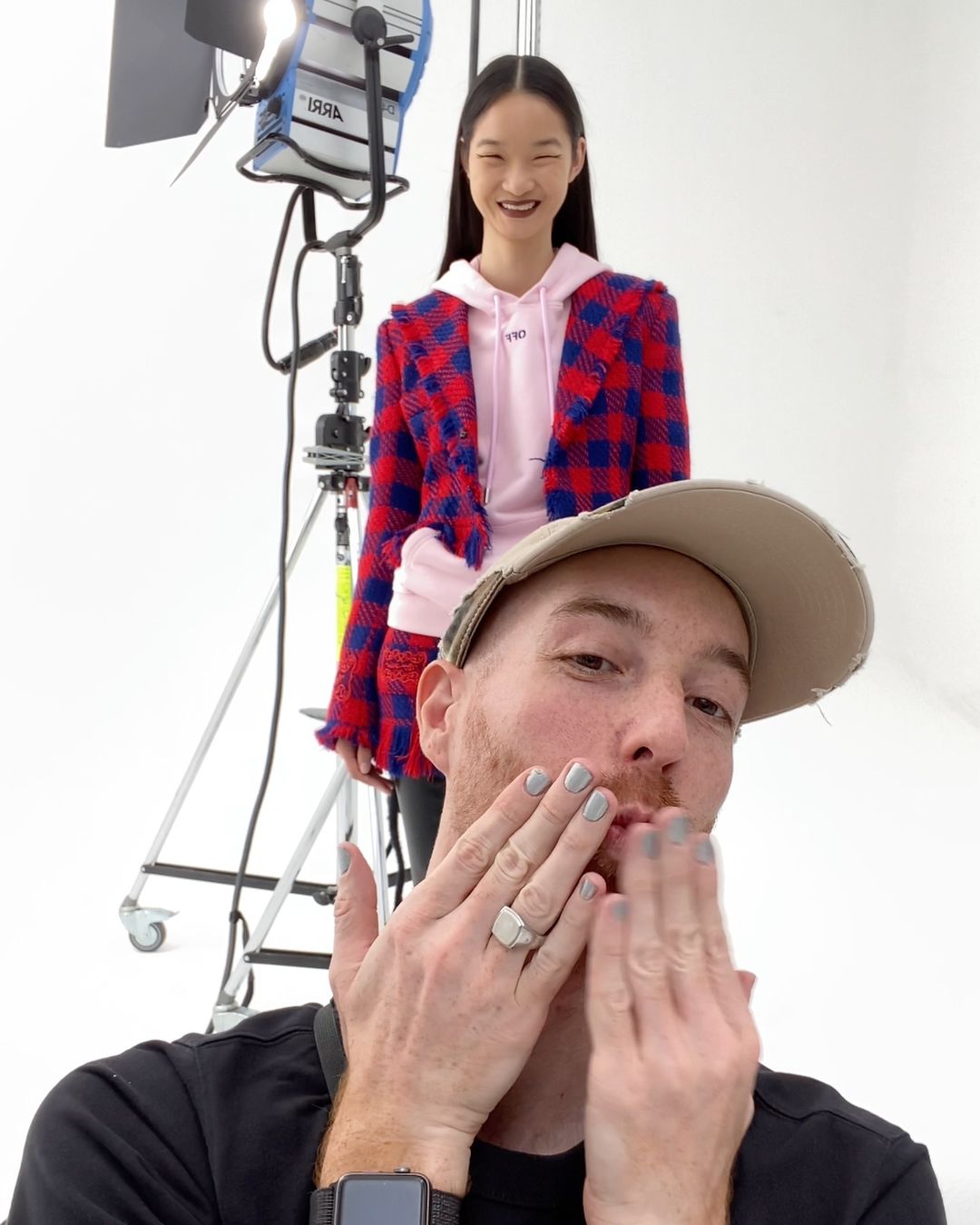Bright studio photo of an Asian fashion model in a pink hoodie and plaid jacket, with a content creator in the foreground blowing a kiss.