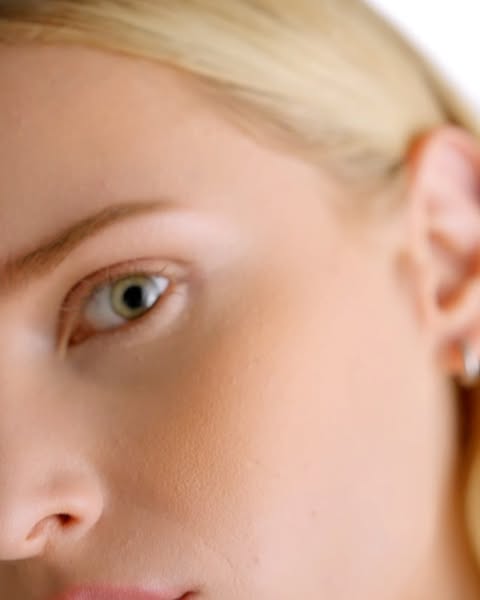 Close-up shot of a fair-skinned adult woman's face featuring blonde hair, an eye, nose, and a glimpse of lips against a plain backdrop.