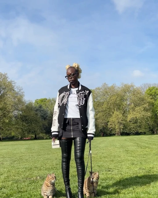 An adult female wearing thigh-high boots and a varsity jacket stands on a lawn in a park with two cats on leashes.