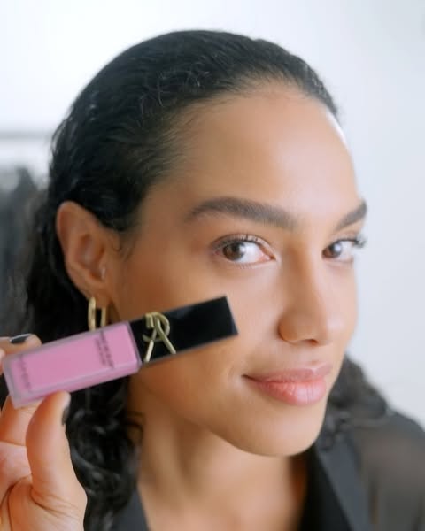 A woman holds a pink lip gloss tube near her face. She has brown skin, dark curly hair, gold hoop earrings, and is wearing a black top.