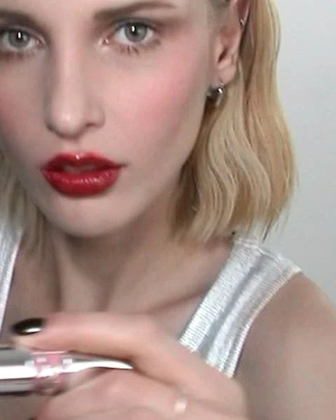 Blonde woman with dramatic makeup and shiny red lips applies a clear cosmetic product with a brush in an indoor setting.