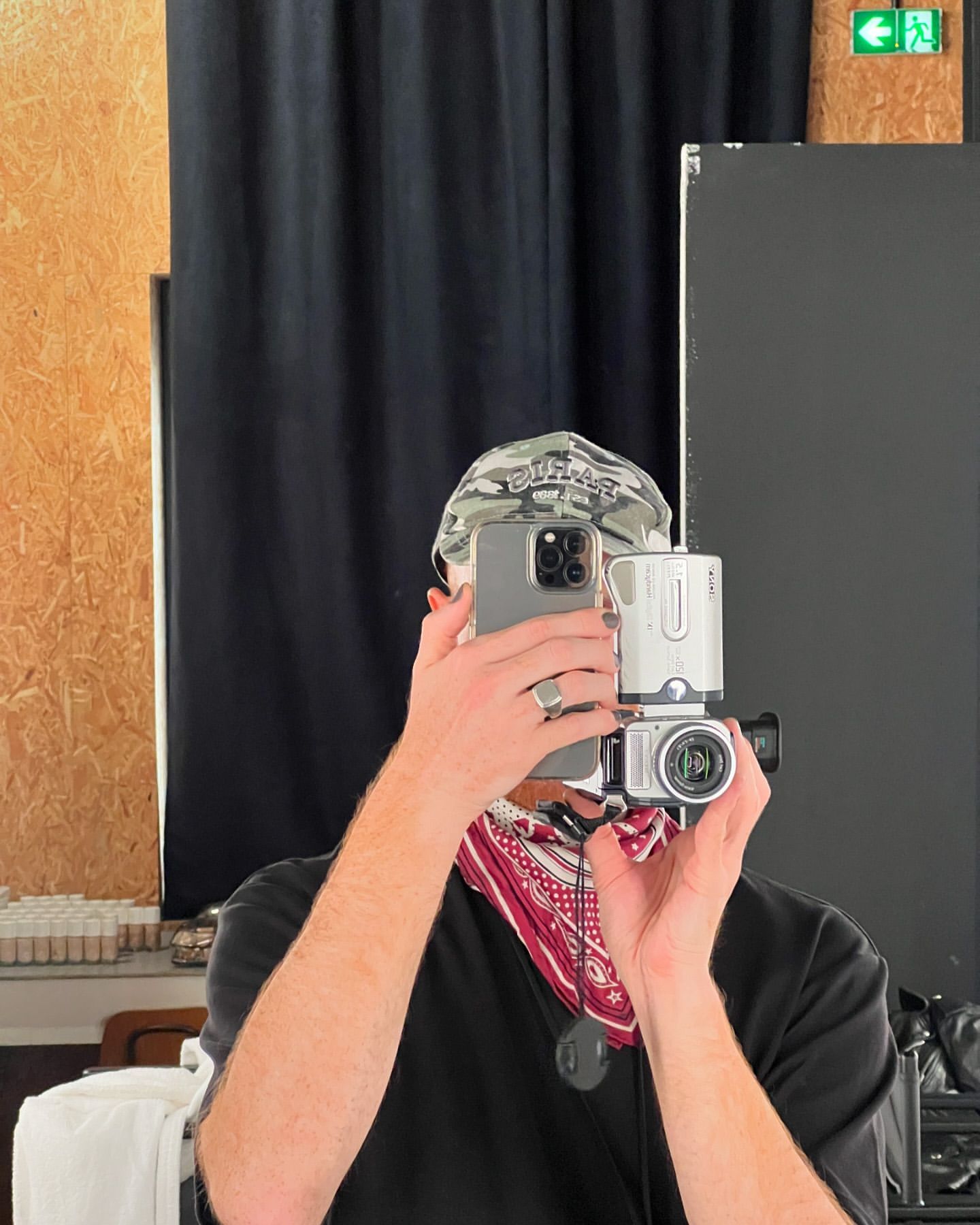 Person in black t-shirt, bandana and cap takes a selfie in a mirror using an iPhone and a vintage camcorder in an indoor setting.