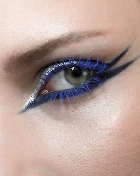 Close-up of a woman's eye with artistic makeup, featuring blue mascara, silver glitter eyeshadow, and dark eyeliner.