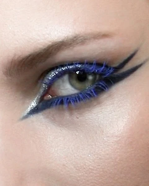 Close-up of a woman's eye with artistic makeup, featuring blue mascara, silver glitter eyeshadow, and dark eyeliner.