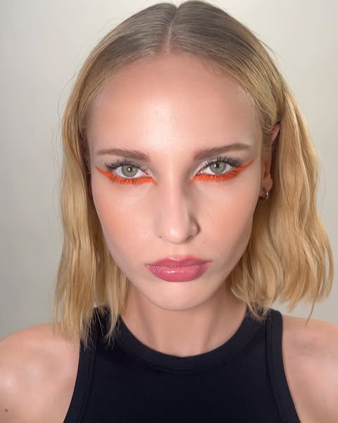 Close-up of a woman with fair skin, blonde hair, and bright orange eyeliner on her upper eyelids. She wears a black top.