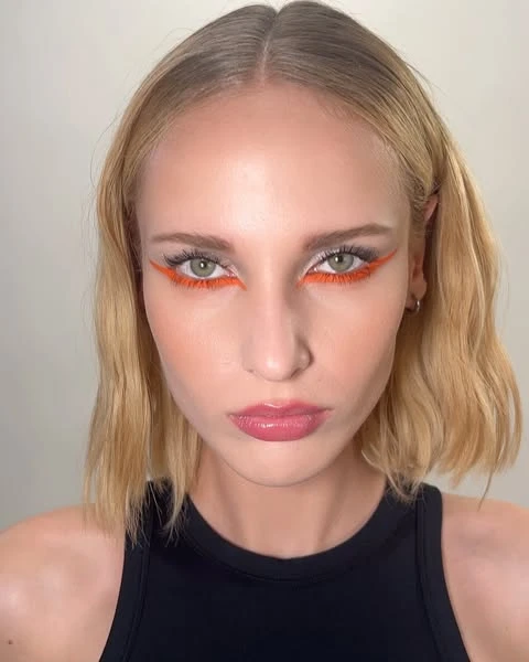 Close-up of a woman with fair skin, blonde hair, and bright orange eyeliner on her upper eyelids. She wears a black top.