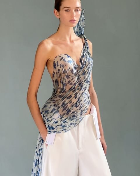 Fashion model posing in a studio wearing a one-shoulder draped top with blue floral print and white pants against a grey background.