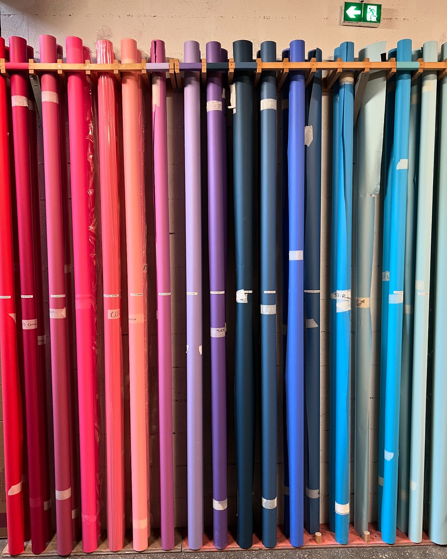 Colorful rolls of paper or fabric are neatly stacked on a wooden rack, displaying a spectrum of hues from red to blue and green.