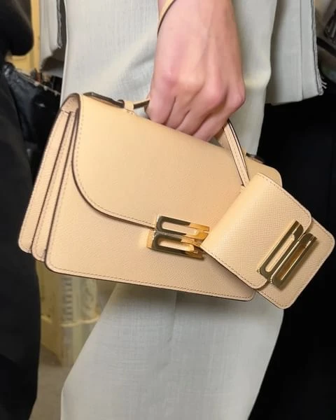 Close-up of a woman's hand holding a beige leather top-handle handbag with gold hardware, paired with a matching card case.