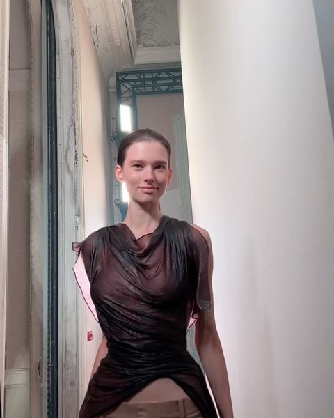 A young adult female fashion model poses indoors wearing a sheer brown top and khaki pants near a window and white column.