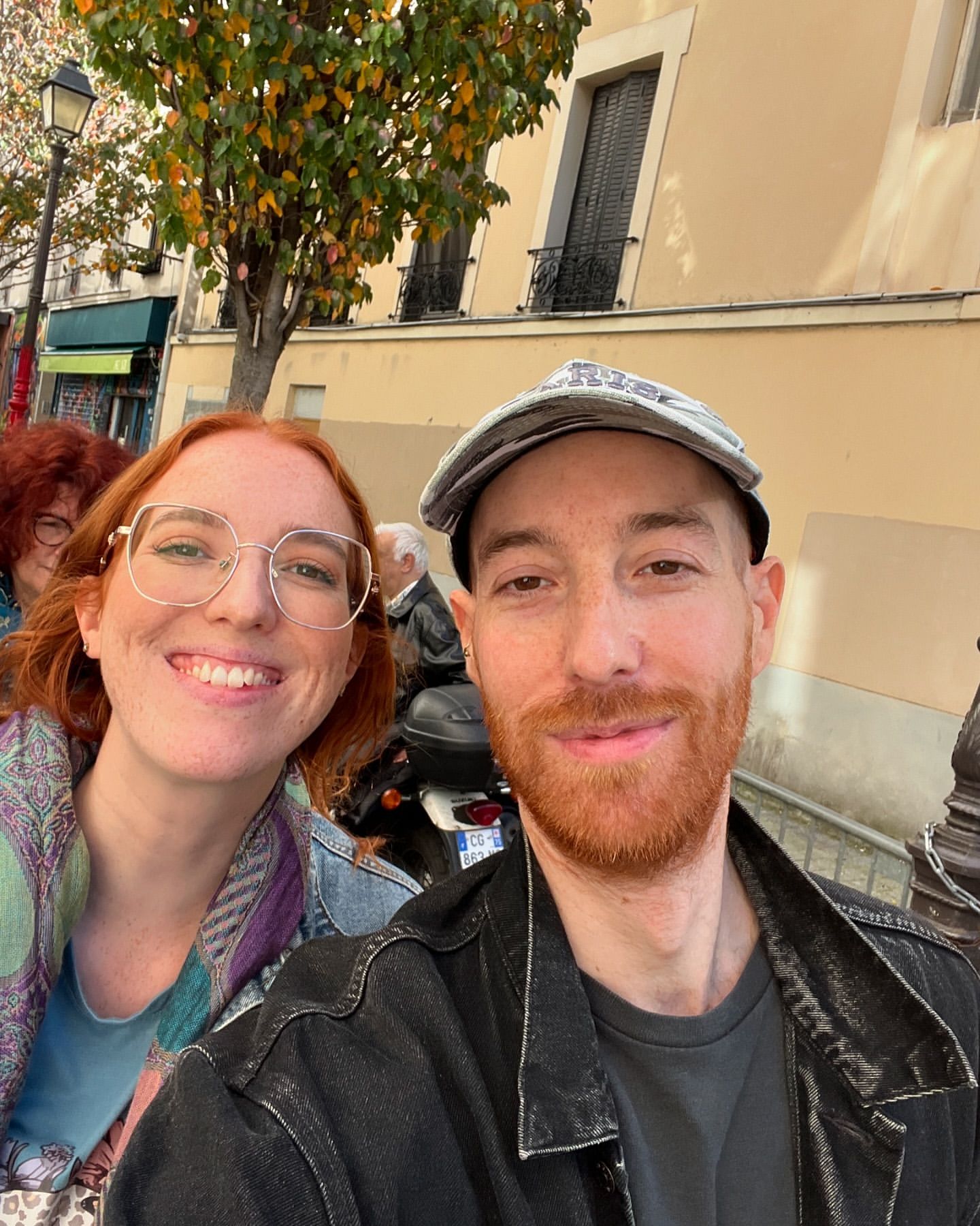 Close-up of a smiling woman with glasses and a cap-wearing man, both with red hair, outdoors in front of a light-colored building.