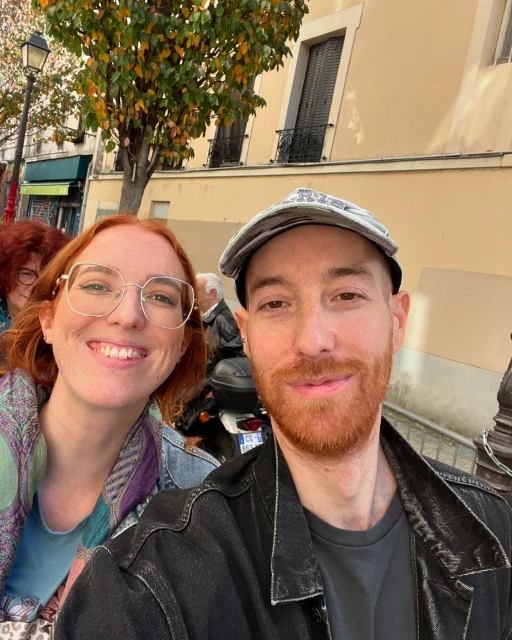 Close-up of a smiling woman with glasses and a cap-wearing man, both with red hair, outdoors in front of a light-colored building.