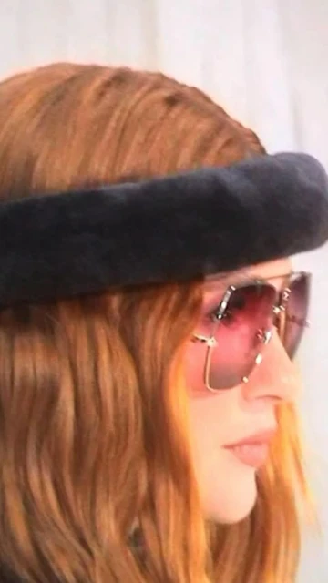 Stylish redhead woman in side profile wearing a fur headband and oversized pink-tinted sunglasses in a close-up indoor portrait.