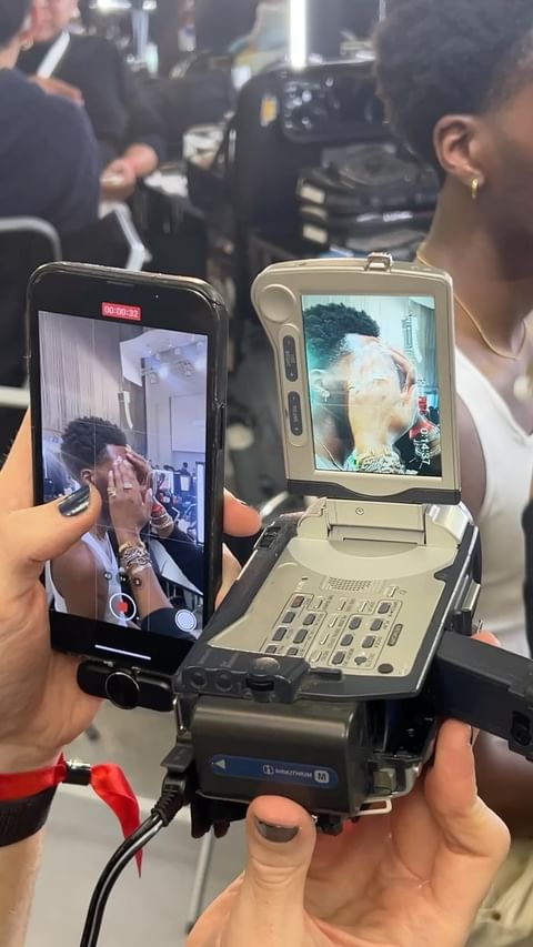 Close-up of a camcorder and phone screen displaying an adult doing makeup; the device is held by a person with a red wrist ribbon.