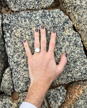 Close-up of an arm and hand with dark gray nail polish and a silver ring lying on a textured granite rock surface.