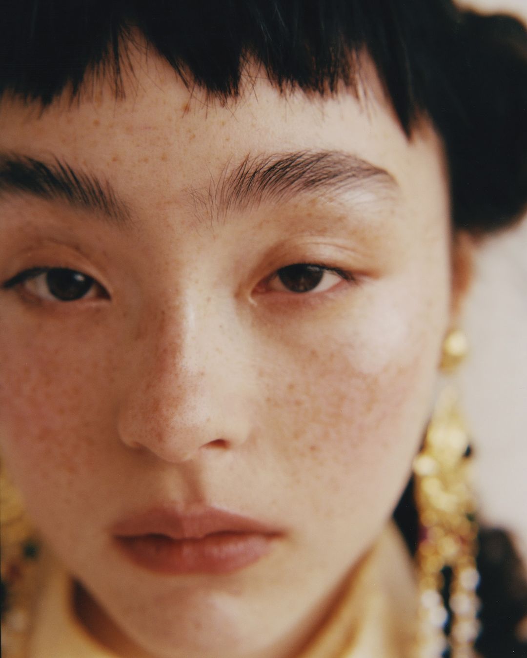 Close-up portrait of an Asian woman with freckles, wearing gold earrings and a yellow top, against an off-white backdrop.