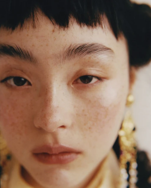 Close-up portrait of an Asian woman with freckles, wearing gold earrings and a yellow top, against an off-white backdrop.