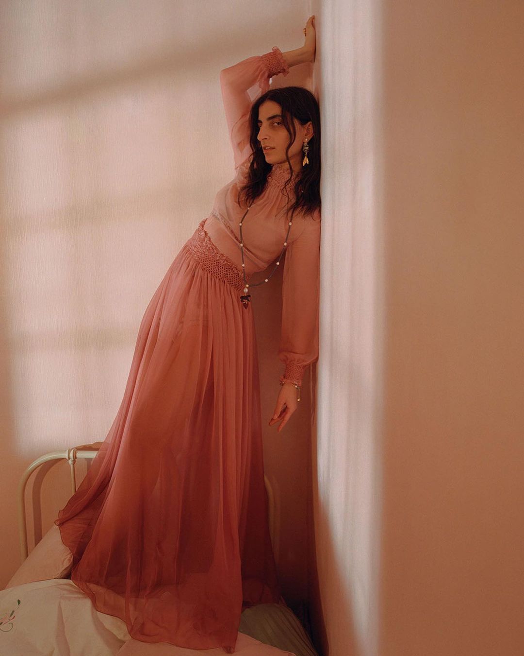 A woman with dark hair in a long pink dress leans against a pink wall in a portrait style with soft light, creating a gentle, ethereal mood.