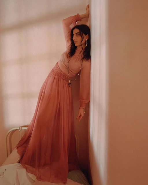 A woman with dark hair in a long pink dress leans against a pink wall in a portrait style with soft light, creating a gentle, ethereal mood.