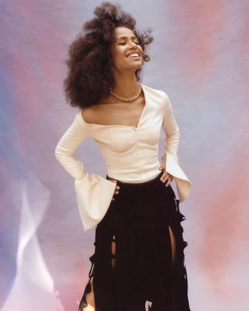 Smiling woman with afro hairstyle wearing a cream-colored top and a black skirt against a mottled pink and purple backdrop.