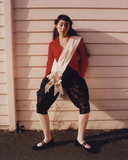 Young woman in red jacket and black bloomers poses against a weathered, light-colored wooden wall in full outdoor sunlight.