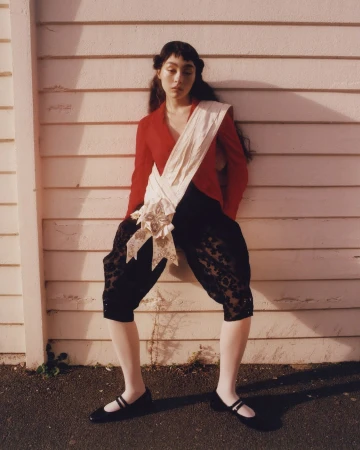Young woman in red jacket and black bloomers poses against a weathered, light-colored wooden wall in full outdoor sunlight.