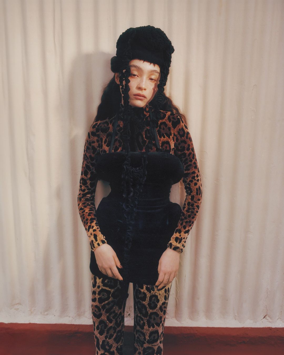 Portrait of a young woman wearing a black fur hat, a black velvet top, and leopard-print leggings posing in front of a white curtain.