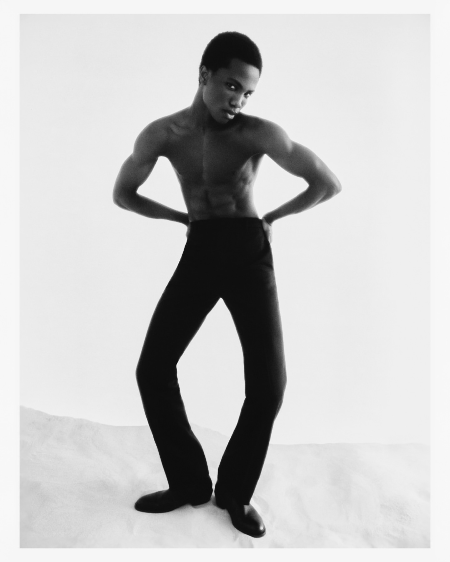Striking black and white studio shot of a slender, shirtless Black male model in flared black trousers, hands on hips, posing dynamically.