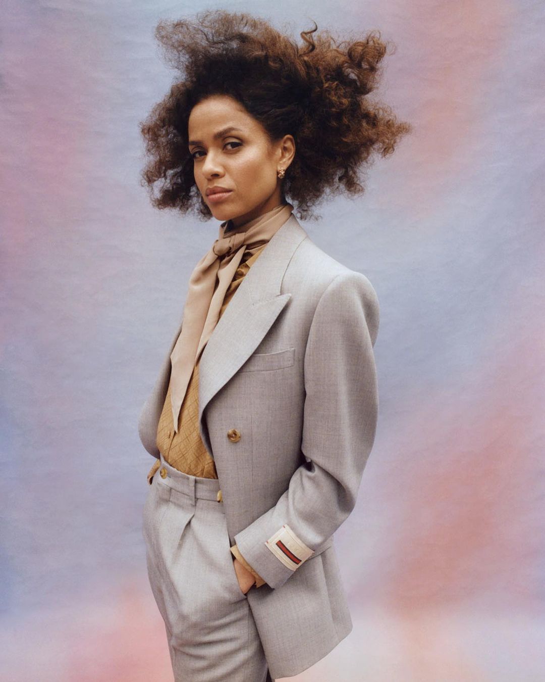 Portrait of a curly-haired woman in a grey pantsuit with a scarf, posing against a gradient backdrop of pink, blue, and peach hues.
