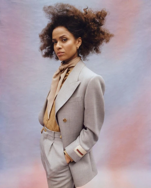 Portrait of a curly-haired woman in a grey pantsuit with a scarf, posing against a gradient backdrop of pink, blue, and peach hues.