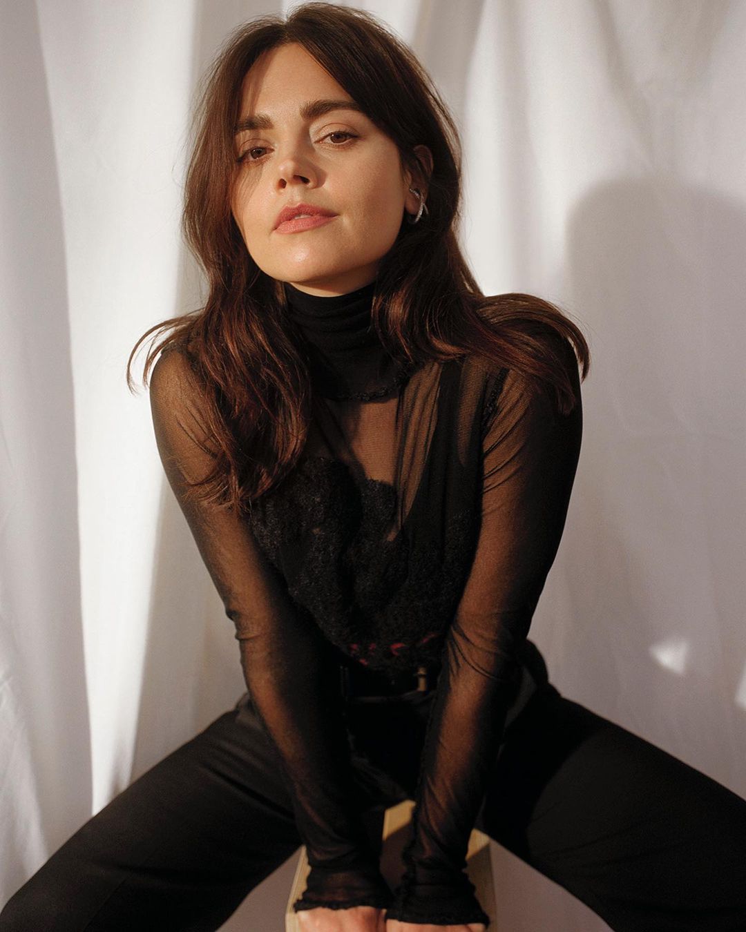 Portrait of a brunette woman with dark sheer top sitting on a stool in front of a white fabric backdrop, casual pose, studio lighting.