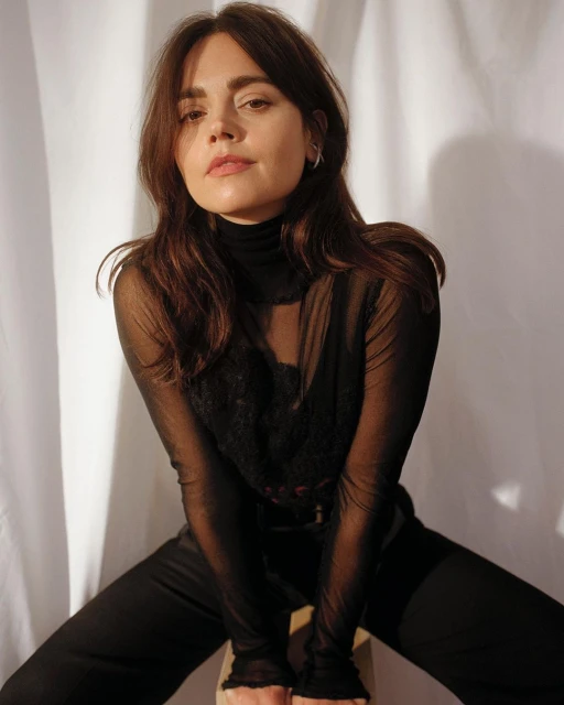 Portrait of a brunette woman with dark sheer top sitting on a stool in front of a white fabric backdrop, casual pose, studio lighting.