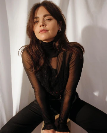 Portrait of a brunette woman with dark sheer top sitting on a stool in front of a white fabric backdrop, casual pose, studio lighting.