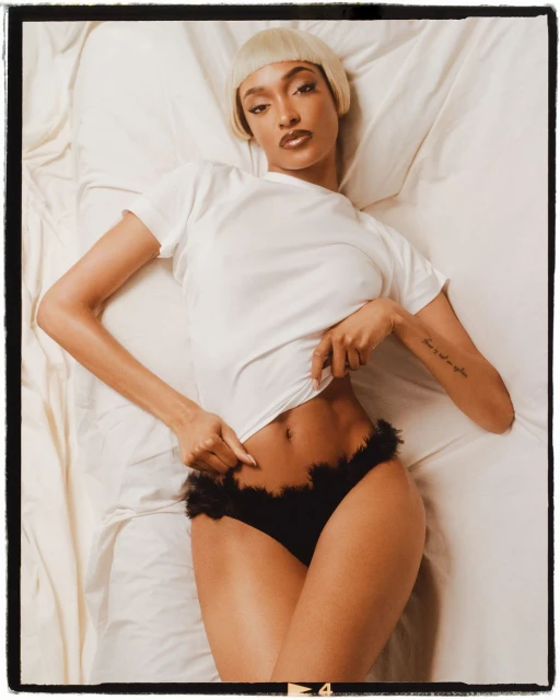 Woman in underwear poses on bed, wearing a white t-shirt and blonde wig, captured in a medium shot with a black border.