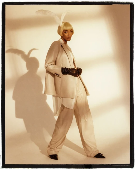 Woman poses in a beige suit and black gloves, wearing a feathered headpiece. Studio lighting and a neutral background.