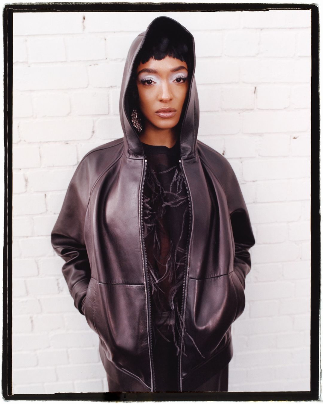 Portrait of a woman in a brown hooded jacket against a white brick wall, featuring dramatic makeup and large earrings. Fashion shot.