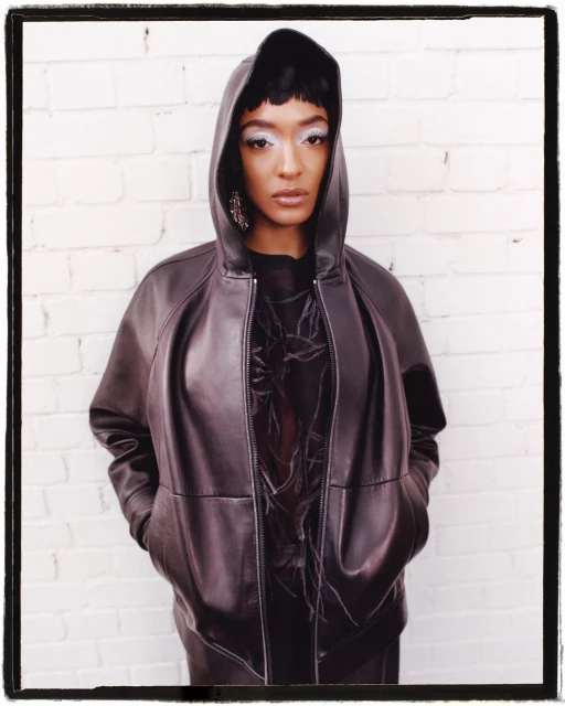 Portrait of a woman in a brown hooded jacket against a white brick wall, featuring dramatic makeup and large earrings. Fashion shot.