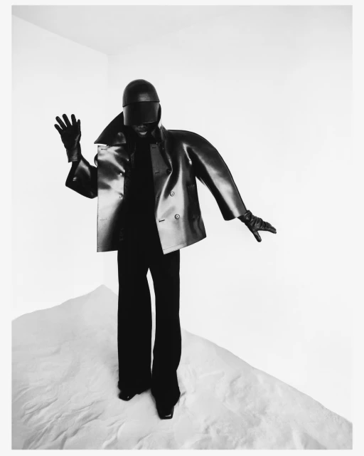 Black and white fashion shot of a person in a futuristic helmet and dark, structured jacket standing on a textured white surface.