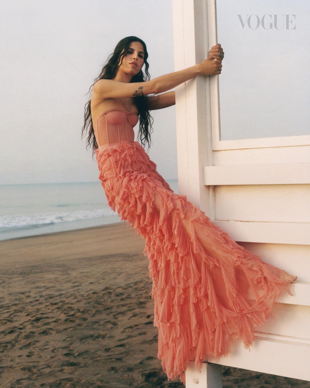 Brunette woman in pink dress leaning against white lifeguard tower on beach. Ocean and sky visible in background. Vogue logo in corner.
