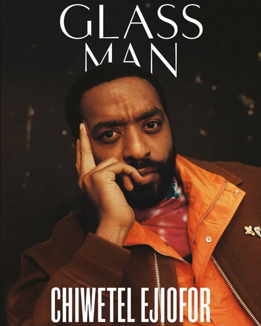 Close-up portrait of actor Chiwetel Ejiofor in brown coat and orange zip-up with "Glass Man" magazine title. Dark, textured background.