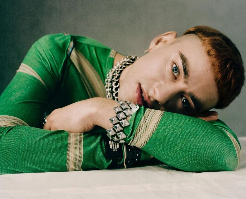 Close-up of a young adult male with light skin, short reddish-brown hair, and striking blue eyes, wearing green striped clothing and silver jewellery.