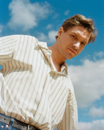 Man wearing a striped shirt posing against a bright blue sky with soft white clouds