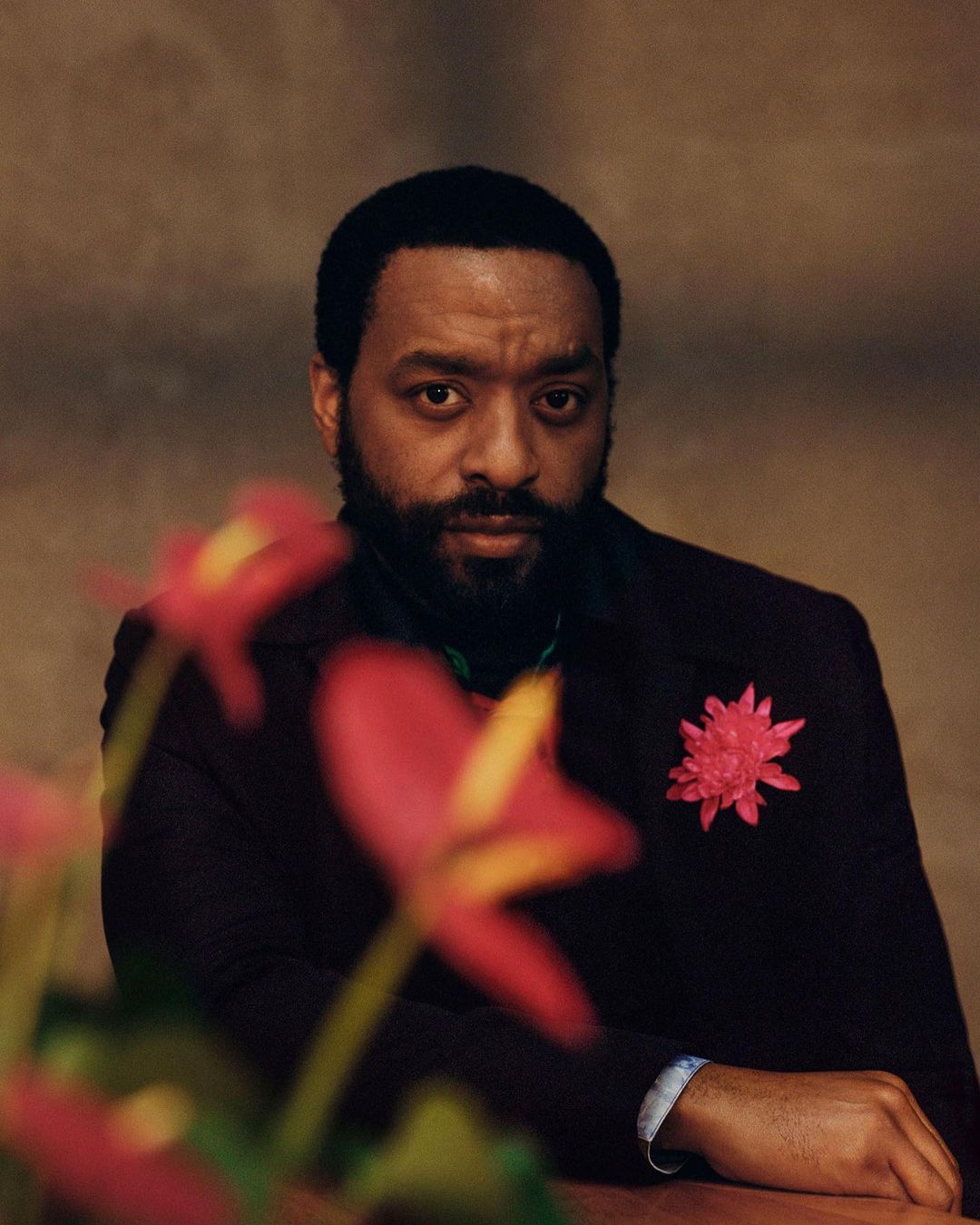 Portrait of Black adult male in a dark jacket with pink flower against a textured background, gazing forward, with flowers in foreground.