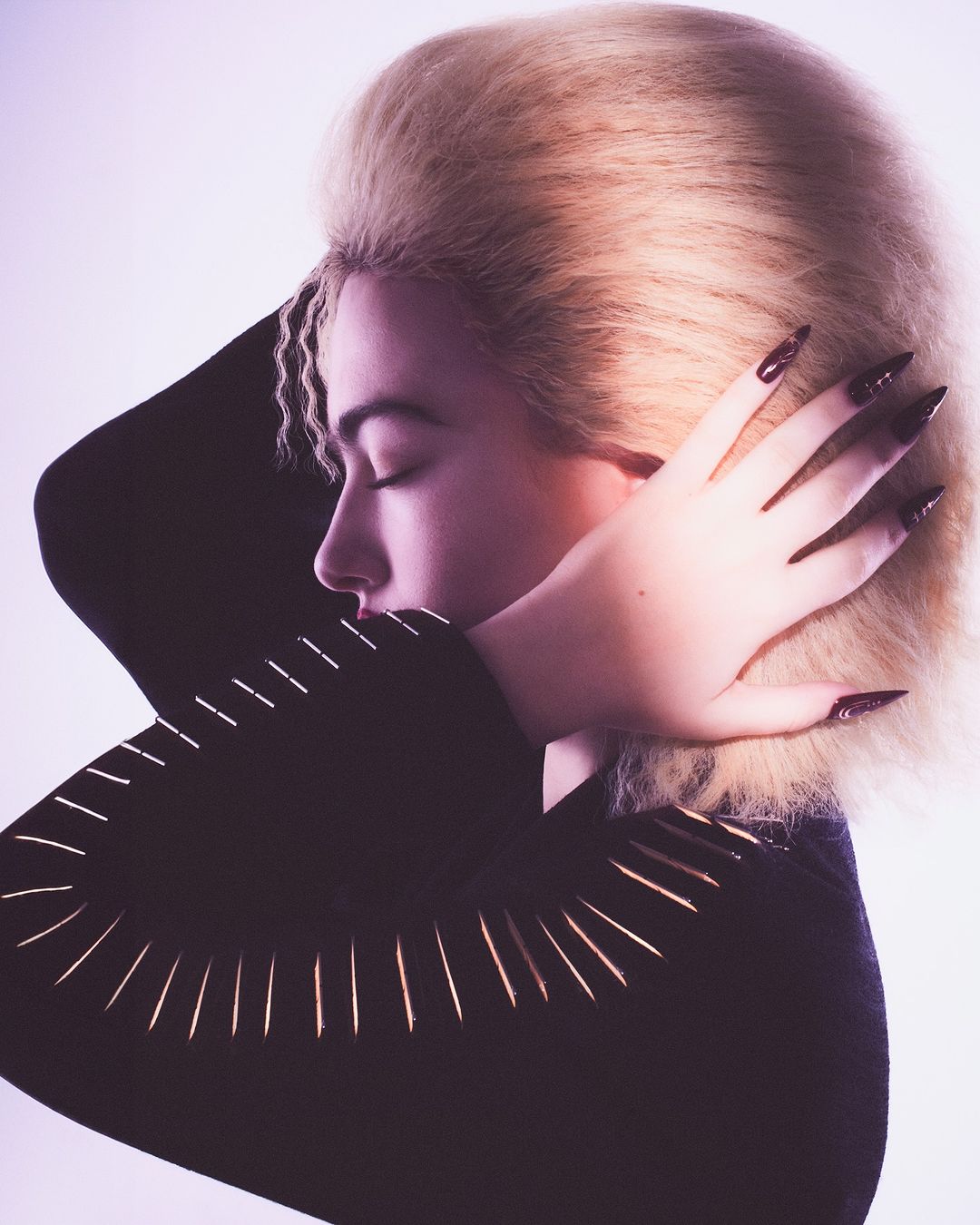 Close-up portrait of a woman with blonde bouffant hair, long dark nails, eyes closed, wearing a black top with gold accents, against a light background.